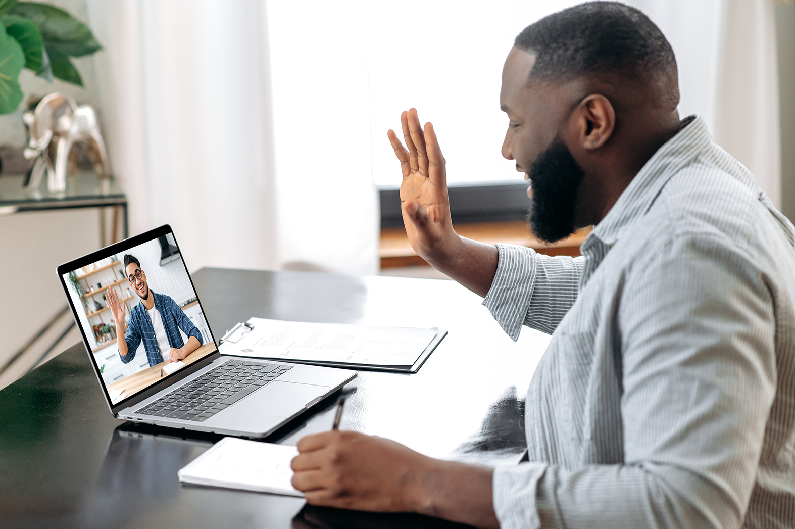 Online video meeting. African American young successful man talking on video conference with his colleague, friend or mentor while sitting at the desk in the office, talking about strategy,takes notes Online video meeting. African American young successful man talking on video conference with his colleague, friend or mentor while sitting at the desk in the office, talking about strategy,takes notes